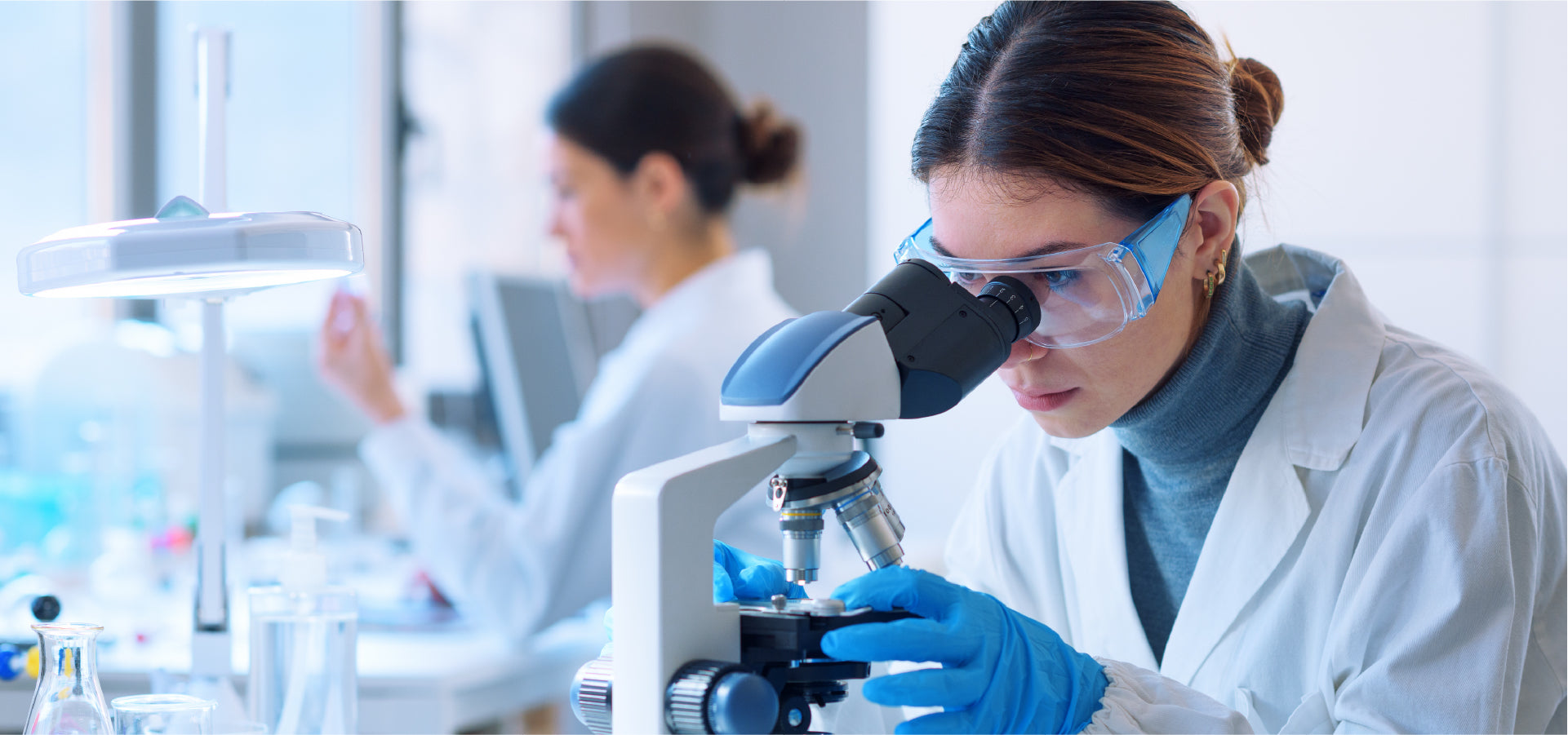 Female scientist wearing safety goggles using a compound microscope in a laboratory.
